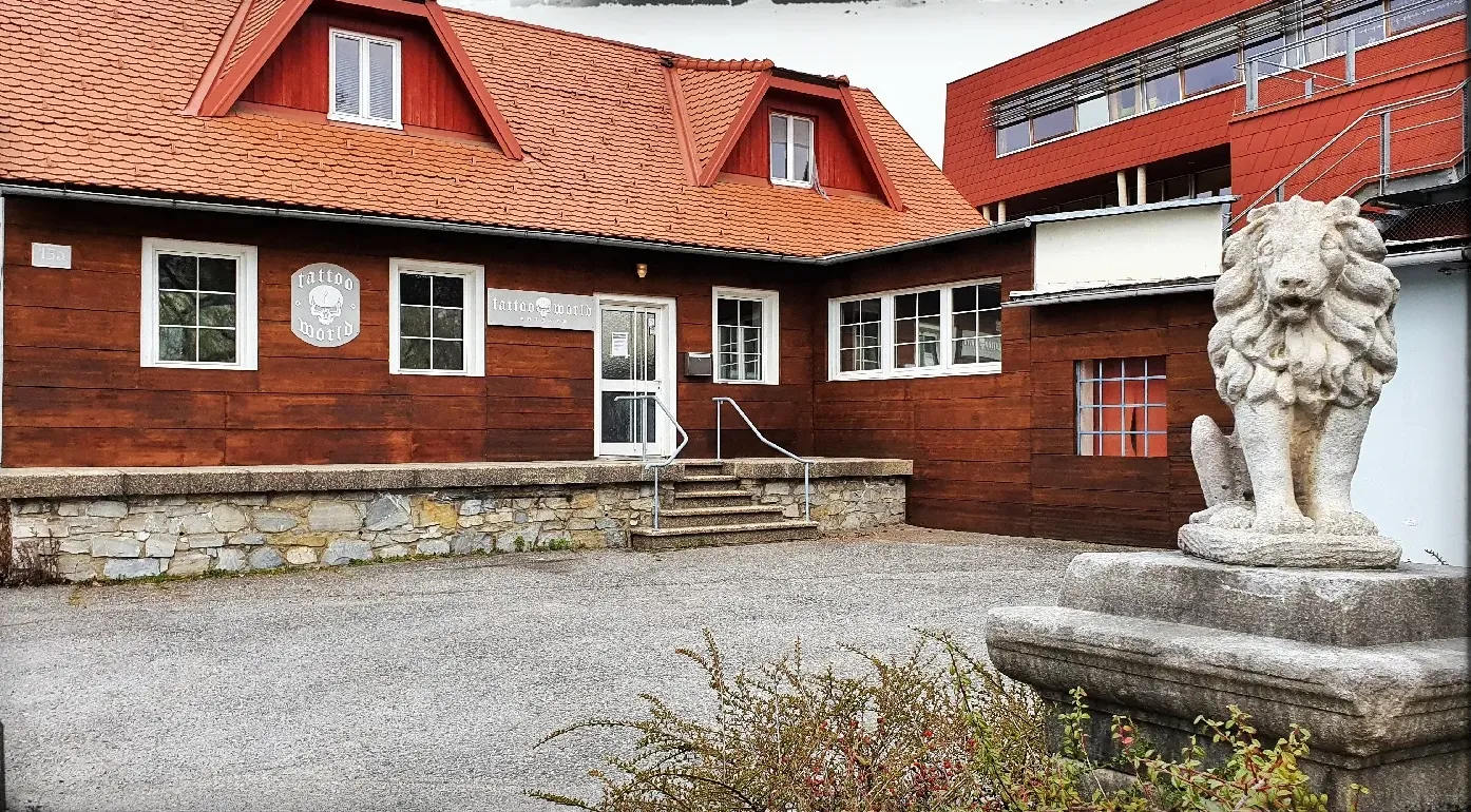 Das Studio: ein rotes Holzhaus mit Ziegeldach, Tattoo-World-Schild und einem Steinlöwen im Hof